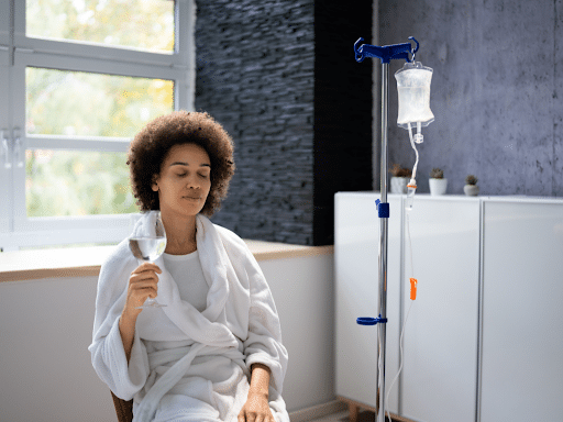 Woman in a robe sitting beside an IV drip stand, relaxing and holding a glass of water during an infusion session.